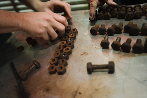 Detail shot showing two hands at the right, arranging two rows of rusted nuts. Another hand extends from the top of the image, to rest on a row of bolts.