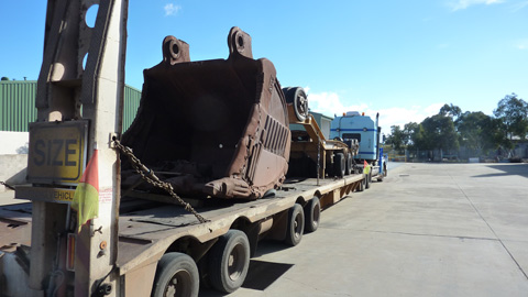 A close up view of a semi-trailer parked on a large open concreted area. The rear of the trailer is closest to the camera, at the right-hand side of the image. The prime mover can be seen at the front of the trailer, in the middle distance of the image. On the tray of the semi-trailer is the large metal bucket from an excavator used in an iron ore mine. The bucket sits facing the rear of the trailer. There is some other mining equipment on the trailer, between the bucket and the prime mover. The bucket is a dark earth colour, stained by its use in the mine. In the left background can be seen part of a large green industrial shed. In the right background of the image can be seen some trees. The sky above is mostly clear of clouds.