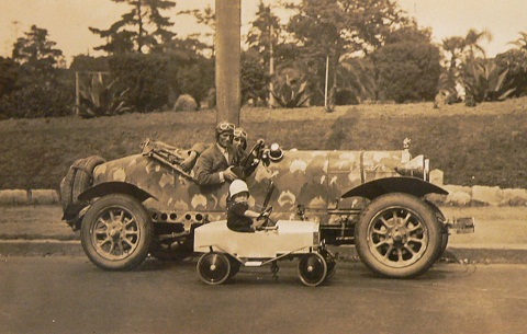 Black and white photo showing a side view of two men wearing leather flying style caps and goggles, sitting in an open-topped car which has small maps of Australia painted on the chassis. Beside them is a small child sitting in an open-topped toy car.