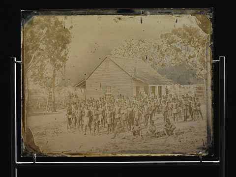 A large group of men, mainly wearing loin cloths, standing and sitting outside a small wooden building, with gum trees in the background.