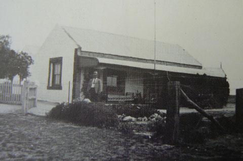 Black and white photo showing a man standing at the front of the small homestead, with a picket fence to the left and a fenced garden at the front. A daybed and wooden chair are on the verandah.