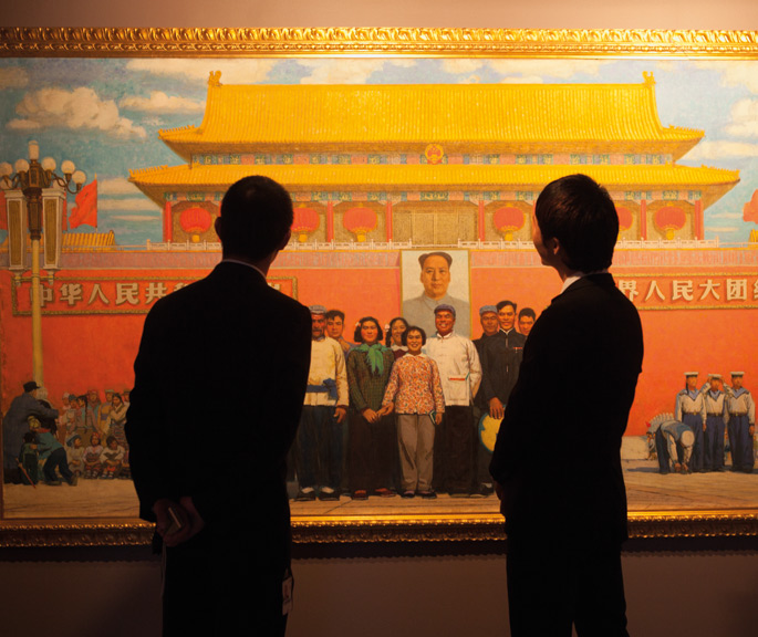 A colour image of two people dressed in dark suits viewing a large painting mounted on a wall.