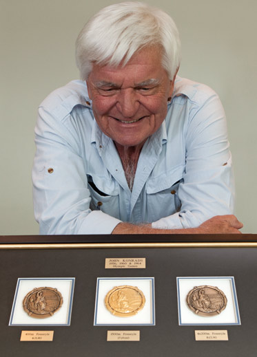 Colour photograph of a man resting with folded arms on a table. In front is a framed set of three circular medals.