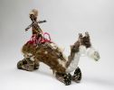 A white, grey and brown model camel with two riders made from, raffia, minarri (greybeard) grass, wool and leather. The two riders are seated on a red crocheted saddle, which is fastened to the camel with leather straps and leads. There are two separate accompanying brown crocheted swags. A 'Tjampi / desert / weavers' label is attached to one foot.