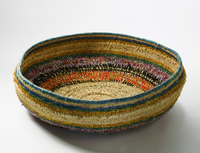 A large shallow basket made of bands of grasses which have been bound in plant material dyed in different colours and in strands of wool.