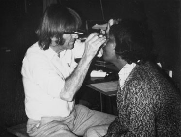 A mean wearing a scope over this eyes sits peering into the eyes of a woman, who leans forward for examination.