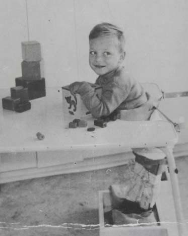 Black and white photo of a young boy at a table. He wears callipers on his legs and is playing with a box and blocks.