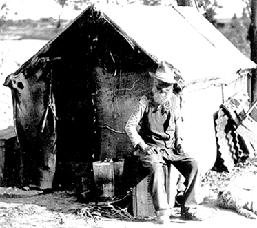 Old man seated on a crate in front of his simple dwelling with a dog at his feet.