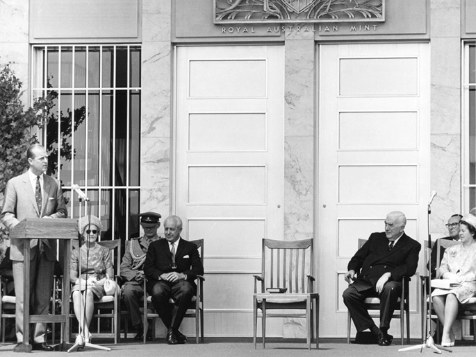 Prince Phillip speaking at a lectern with Holt and Menzies seated. Other dignitaries and their wives are also present. Behind them is the main door of the Mint.