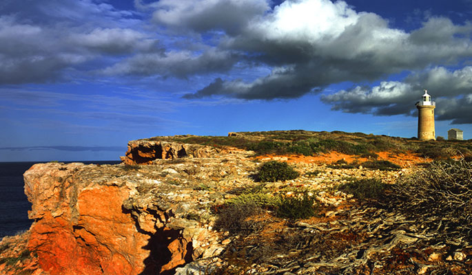 Colour photo of a piece of coastline showing cliffs of orange rock and a small lighthouse in the distance.