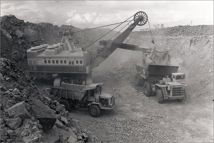 Black and white photo of large power shovel dumping ore into one of two large mining trucks.