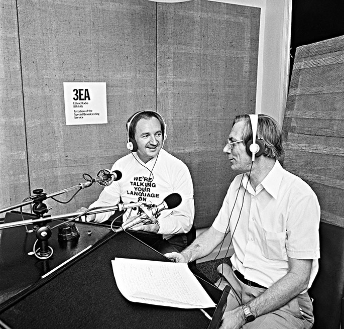 two men sitting in a radio studio in front of microphones. They are smiling and clearly in conversation. The present is wearing a top saying ‘We’re talking your language on 3EA’