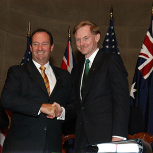 Vaile and Zoellick pose for the cameras shaking hands and smiling. Behind them are a series of Australian and US flags along with a man and a woman smiling and clapping
