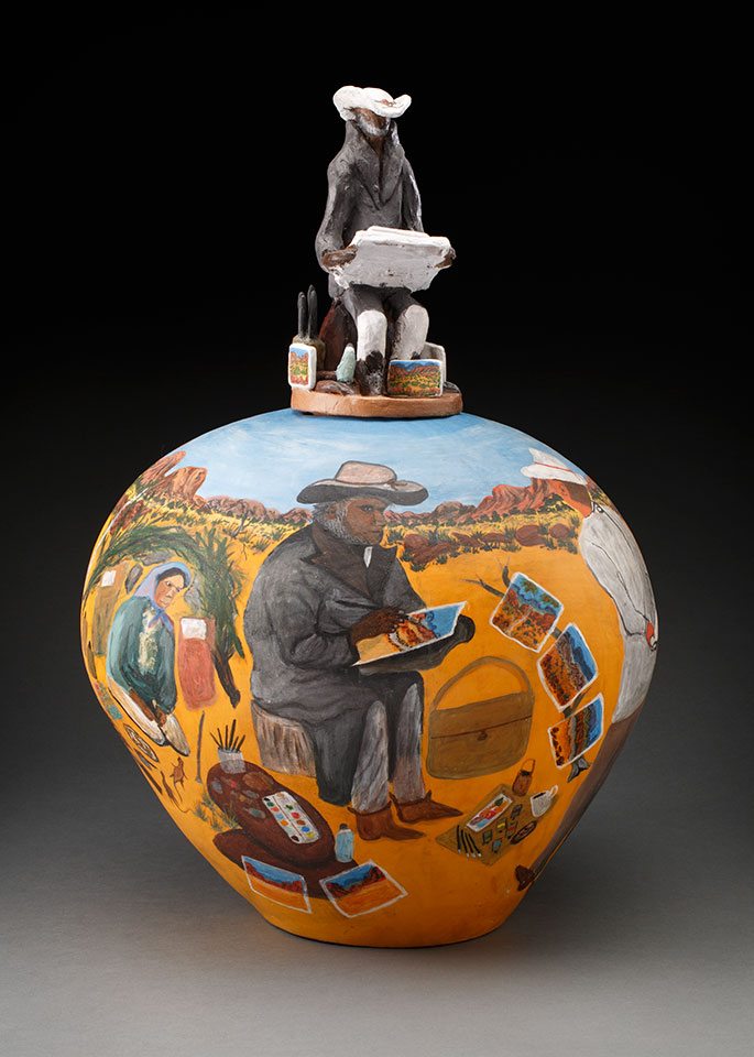 A bulbous pot painted in bright colours, showing a view of an Aboriginal artists at work in the desert. The pot's lid is a sculpted figure of a seated artist.