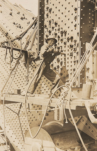 A man stands on a plank, which is attached to the bridge by ropes. He is holding a rivet gun against the metalwork, which is covered in rivets.