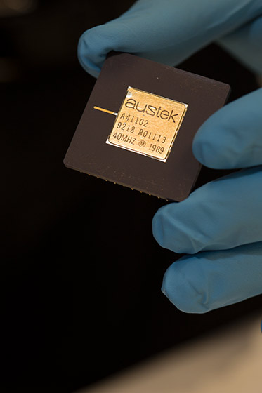 a hand wearing plastic glove holds coin-sized black square with gold-coloured chip at its centre