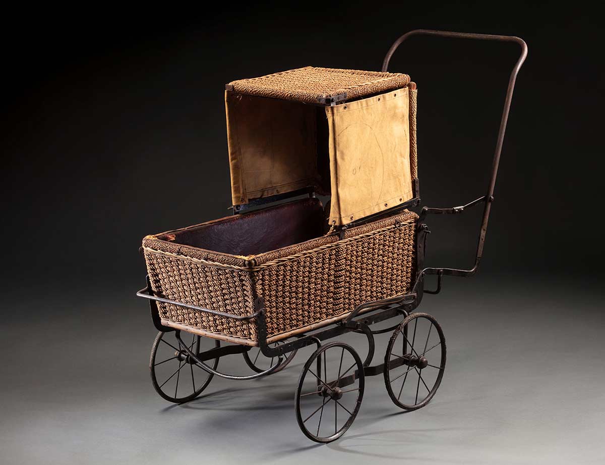 A folding pram (perambulator) with a metal frame and woven wicker body and hood that has separate canvas flaps attached to the left and right side, 1930s.