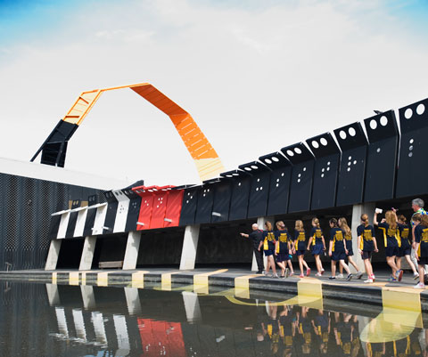 A colour photograph of a group of school children walking through part of the National Museum of Australia. In the foreground is part of a pool of water. The children are walking on a concrete path that runs along the edge of the pool. They all wear yellow and blue uniforms. Behind them is a covered walkway. In the middle distance part of a large curved metal structure can be seen. Beyond that is the sky, which is partly obscured by clouds.