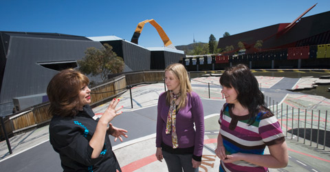 A museum visitor host explains the Garden of Australian Dreams to two visitors. Behind them them is an open area with markings on the ground - a miniature asphalt roadway is painted on the ground and runs diagonally across the image. The Museum's building in the background with the orange loop visible above the roofline.