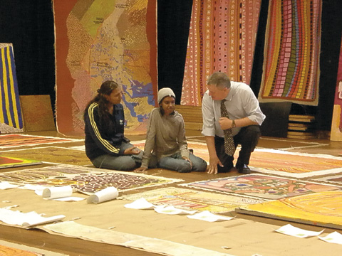 Louise Mengil and Hayley Atkins sit on the floor, beside a kneeling Craddock Morton. The three are surrounded by art works from the Canning Stock Route collection. Several larger canvases hang on the walls in the background.