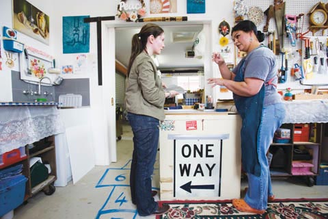 Museum curator Megan Parnell talks with artist Fatima Killeen in a studio. They stand either side of bench where a large 'ONE WAY' traffic sign is taped.