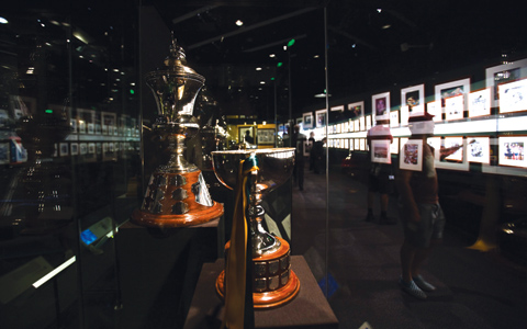 Inside the gallery. Two large trophies in a glass case in close-up on the left side. Brightly lit photographs stretch along a wall into the distance on the right with several people looking at them. The glass of the case reflects the light bouncing off the photographs and partially obscures the people.