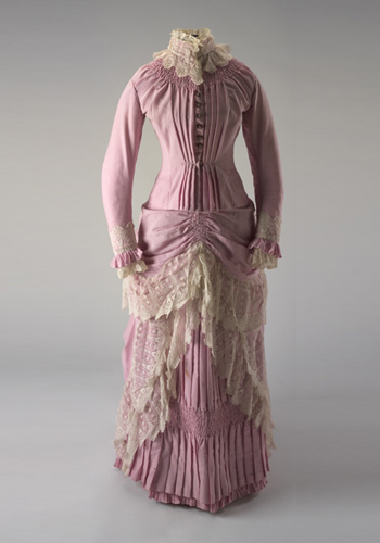 Two piece pink full length wool dress (bodice and skirt) with cream lace trimming. The pleated bodice has a high stand-up collar and is long-sleeved. Both cuffs and collar are trimmed with cream lace. Round copper buttons run down the centre-front and feature an anchor design. The skirt is pleated and layered with extra fabric and trimmed with cream lace.