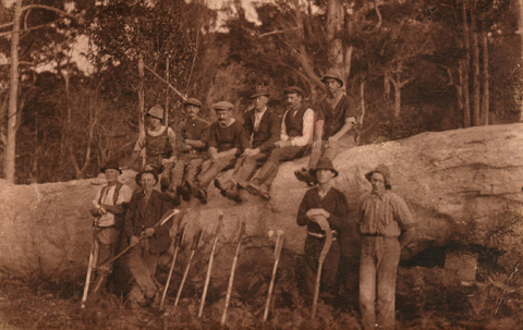 Sepia image showing six men sitting on a felled tree trunk, with four group settlers standing in front. Tools including axes and scrub cutters are propped against the tree.