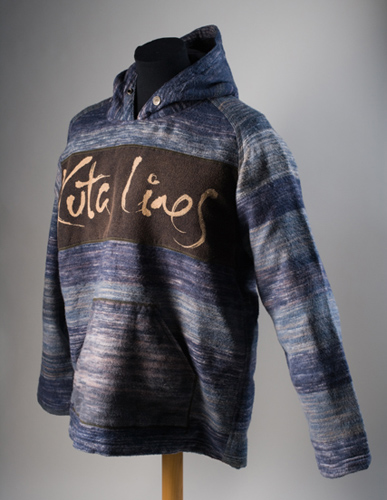 A man's fleecy hooded jumper made from resist-dyed woven cotton fabric of mixed streaks of blue, light blue, black, grey and white. A panel of black fabric at chest-height on the front of the jumper shows the name 'Kuta Lines' in yellow hand-written-style script.