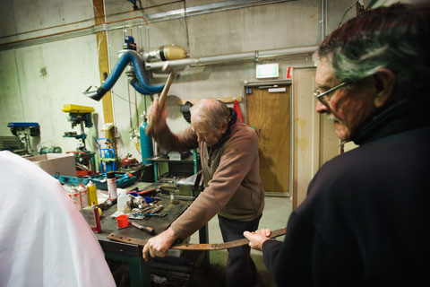 A man standing beside a workbench uses a hammer to strike a thin strip of metal. Another man, seen side on, holds the other end of the strip.