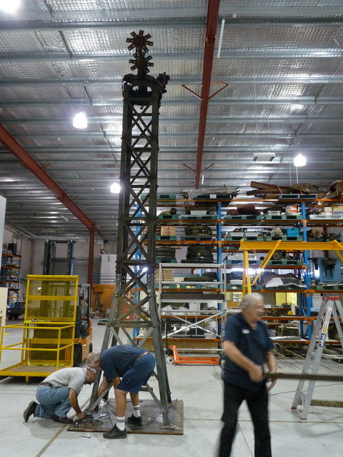 Two men crouch with their backs to the camera, working on a tall steel construction. The metal tower is mounted on a square base plate and rises several metres towards the roof of a workshop. A third man, holding a piece of metal in his right hand, is blurred as he walks away from the construction.