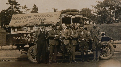 Black and white image showing six men standing in front of a truck which has a canvas cover at the back bearing the words 'BARLOW MOTORS / Melbourne'. All the men wear suits. Those standing second and fourth from left also wear leather flying-style caps and goggles on their heads.