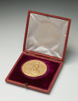 A gold medal in a presentation box. The medal shows an image of a woman holding a wreath and a scroll, with the text 'OB TERRAS RECLUSAS / ROYAL GEOGRAPHICAL SOCIETY OF LONDON'. The case is lined purple velvet, and white satin on the lid.