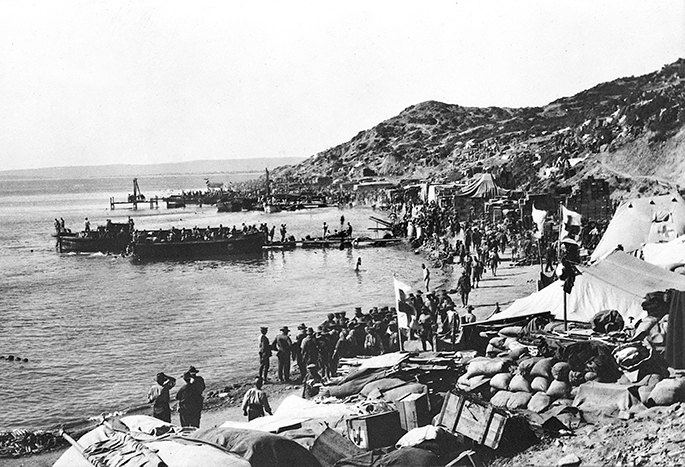 Slightly damaged black and white photo of soldiers on a narrow beach with steep escarpment behind them.