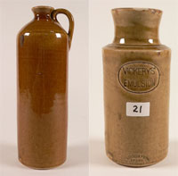 Two tall cylindrical honey brown glazed vessels. On the left is a jug with straight sides tapering to narrow neck and a single handle attached at neck and shoulder. On the right is a jar with a rounded base and straight sides, tapering in to the neck then flaring at the lip. Stamped into one side is the text 'VICKERYS' EMULSION' in an oval frame. Stamped just above the base is the text 'THE BENDIGO POTTERY / EPSOM / BENDIGO' also in an oval frame. A white sticker with '21' handwritten in black appears between the two stamps.