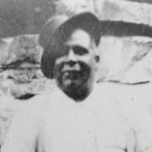 Aboriginal man, smiling, stands facing the camera, a hat set at jaunty angle on his head.