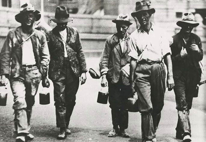 Five men walking towards the camera dressed in shabby clothes. Most are carrying swags and bill cans