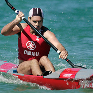 man in red singlet with Yamaha printed on it paddles a surf ski.