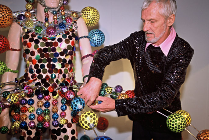 Man in sequined top adjusting colourful adornments on a costume that is worn by a mannequin