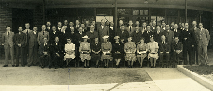 Posed photo of about 20 men standing behind six women sitting.