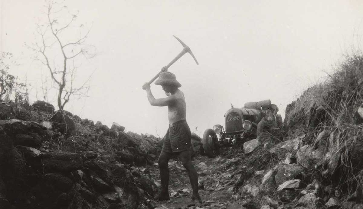 Black and white photo showing a the side view of a topless man wearing a pith helmet, with an axe raised above his right shoulder. He stands down a rocky gorge. An open-topped car sits at the top of the gorge.