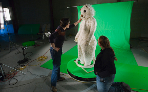 A human-sized white-coloured possum costume on a free-standing frame is posed in front of very bright green background fabric. Two people, one on either side of the costume have their backs to the camera, the person on the left is reaching towards the costume's nose.