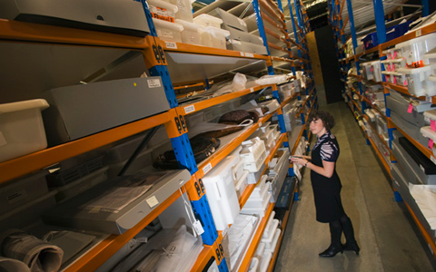 An aisle of tall shelving containing numerous and differently sized boxes. A woman holding a bar code scanner stands in the foreground.