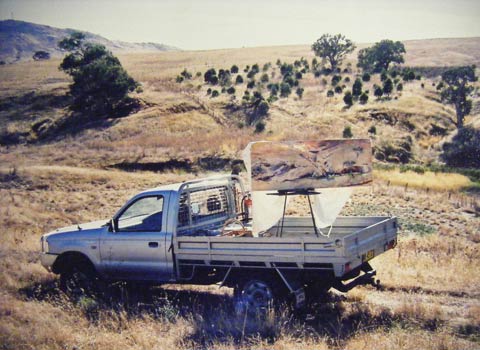 A silver utility vehicle with a tray back is parked in front of a creek bed, with trees and grasses in the distance. An easel supporting a landscape painting sits on the back of the ute.