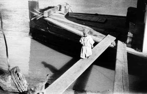 Black and white photo of a young child walking along a gangplank, following a cat.