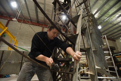 A man stands under a large metal structure. He holds a rubber mallet in one hand and a piece of galvanised steel in place with the other.