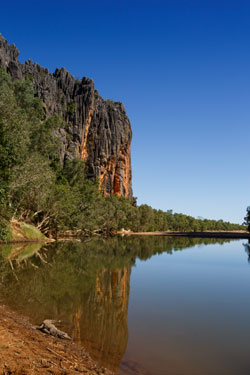 A large expanse of water in front of a large cliff. A crocodile is in the foreground.