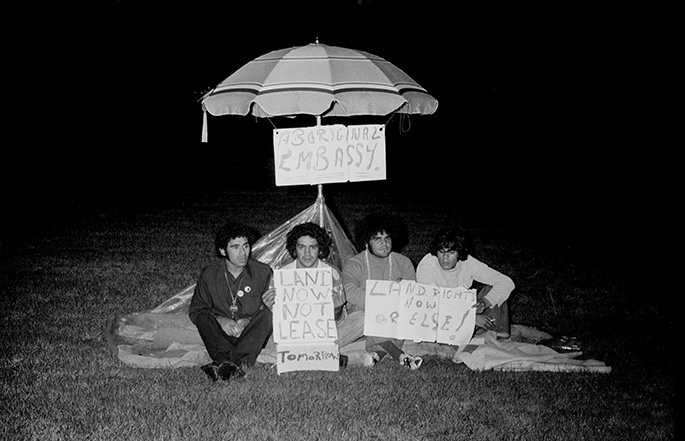 four young men sit in front of a beach umbrella at night holding up signs
