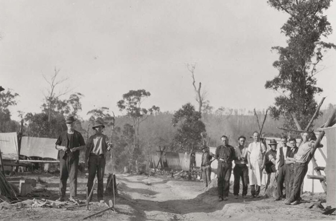 A group of 12 or so men in work clothes pose for the camera. They are in the bush on either side of a dirt track and surrounded by what appear to be tents. It’s possible they’re involved in land clearing.