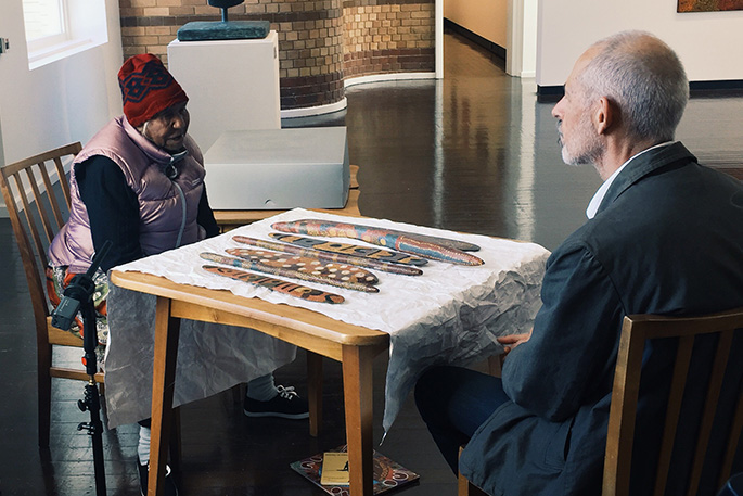 Tilau Nangala and Senior Curator Peter Thorley sitting at a table which has Aboriginal artworks laid on top of it.